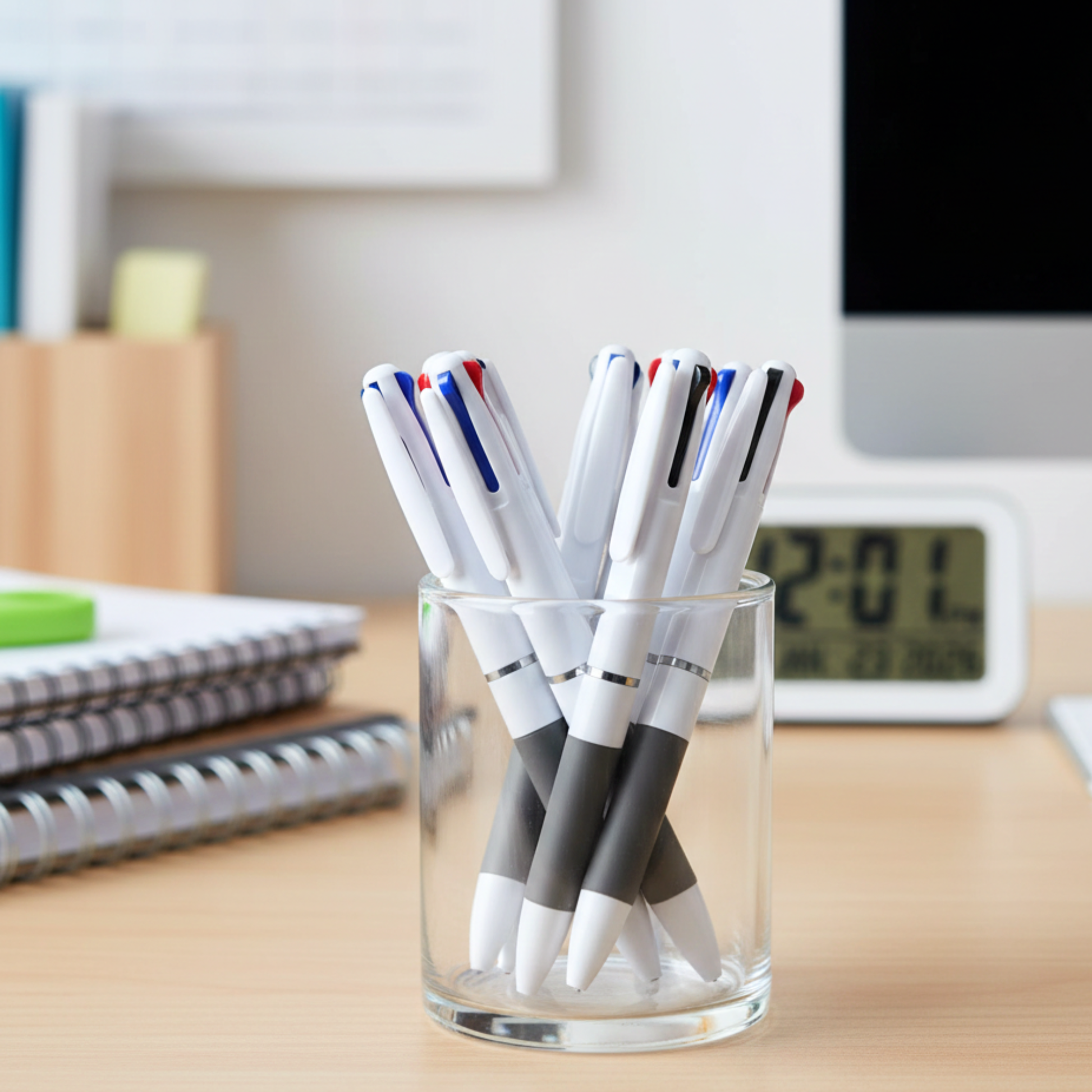 Clear glass pen holder with pens on a desk next to a computer monitor and alarm clock.