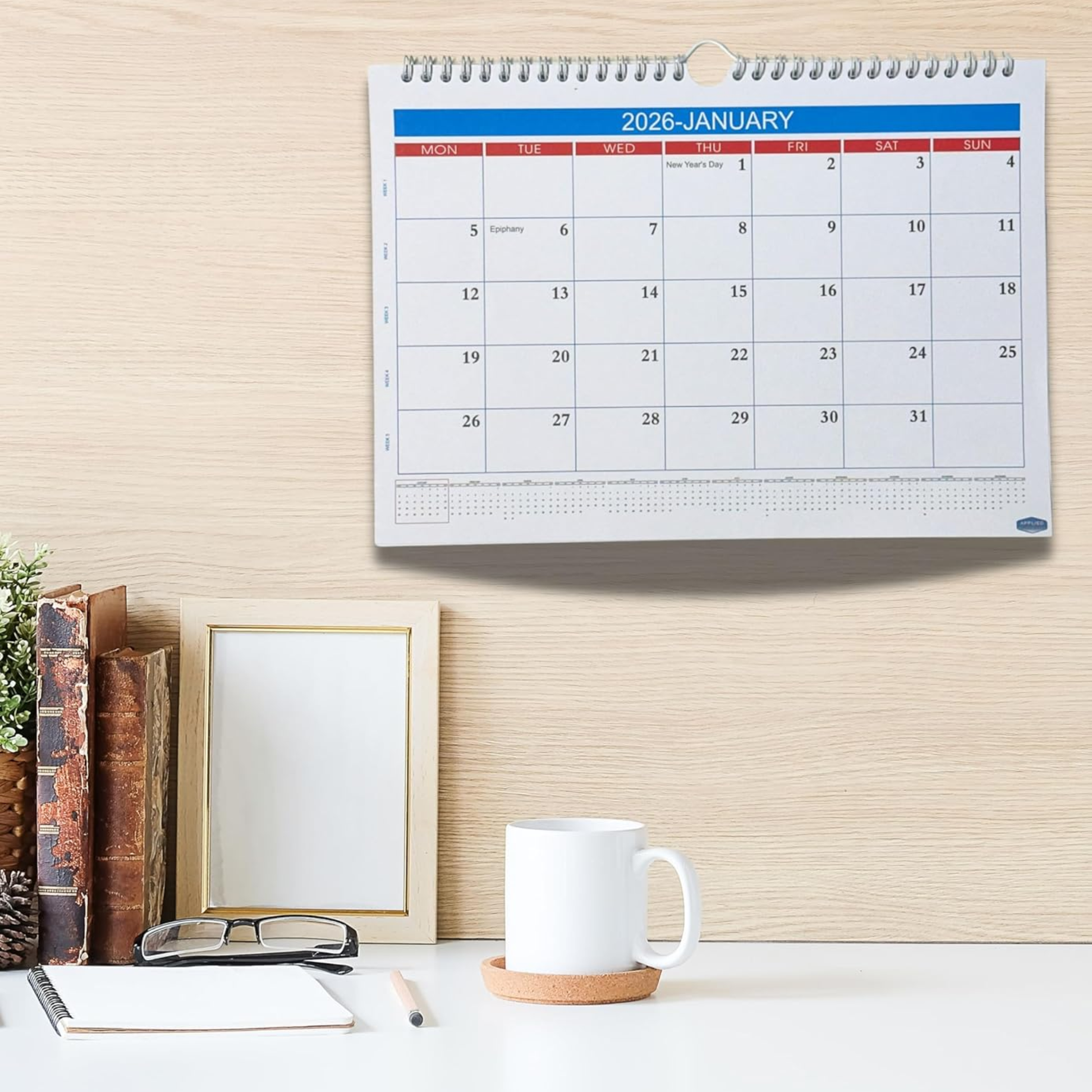 Calendar on a wall with a cup, books, and glasses on a desk below.