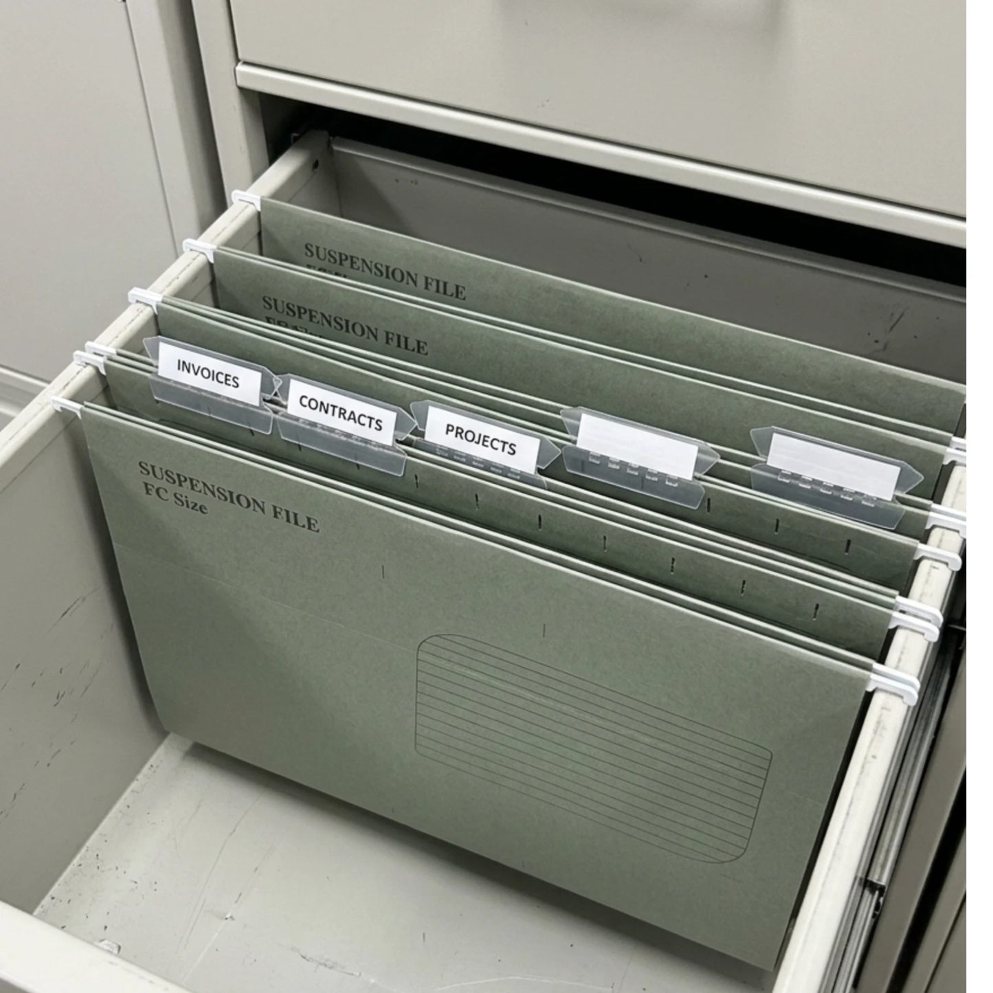 A set of hanging file organizers in a muted sage green color, pulled out of a drawer. The folders are labeled for suspension files and include tabs for invoices, contracts, and projects.