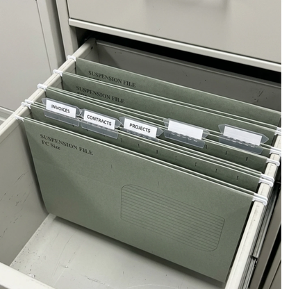 A set of hanging file organizers in a muted sage green color, pulled out of a drawer. The folders are labeled for suspension files and include tabs for invoices, contracts, and projects.