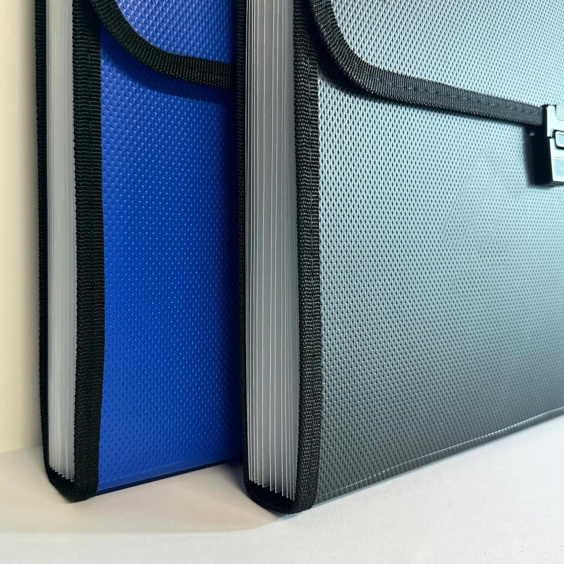 Two textured expanding file or folders shown close up on a light surface. One is blue with black trim, and the other is dark grey with black trim and a small metal clasp visible on the right.