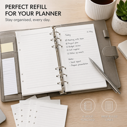 An open gray leather planner ring binder on a wooden desk, showing lined pages with handwritten notes. A pen rests on the right side, and small desk accessories including a mug, plant, and loose page inserts are arranged around it.