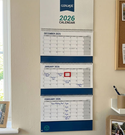 A Concept 2026 wall calendar hanging on a beige wall, showing the months from December 2025 to February 2026 with handwritten notes in blue and a red highlight on January 1st.