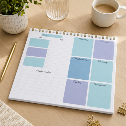 A spiral-bound planner page with pastel weekly and habit tracker stickers displayed on a wooden desk, surrounded by a plant, glass, coffee cup, pen, and binder clips.