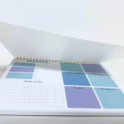 An open spiral-bound planner or agenda showing printed weekly and daily layout stickers in pastel blue, purple, and teal on white paper.